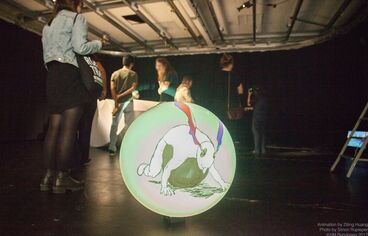Hassenberg Fotografie eines Ausstellungsraumes. Auf dem Boden befindet sich eine runde Scheibe, auf welche das Werk „Hassenberg” abgebildet ist. Zu sehen ist eine Person die mit ihrem Bauch auf einer Art Ball liegt und aus den Ohren der Person steigt bunter Rauch auf.