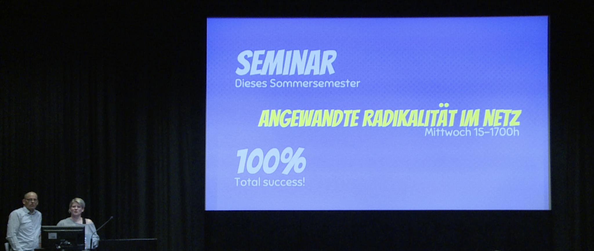 Fotografie der Antrittsvorlesung von dem Künstlerduo Ubermorgen, in der unten linken Ecke sind Prof. Hans Bernhard und Prof. Liz Haas zu sehen. Außerdem eine Projektion in der Mitte des Bildes mit dem Titel der Veranstaltung: Angewandte Radikalität im Netz