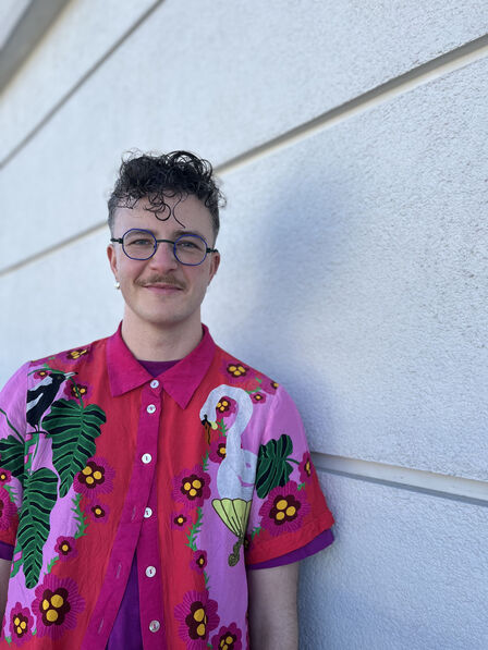 Ren looks at the camera outside in front of a white brick wall they are a white passing trans masculine person with a new mustache and purple octagonal glasses. They are wearing a button down shirt with a kookaburra and a goose in a bathtub on it amidst other flowers and plants. In their ears are earrings and their hair is brown, short and curly. 