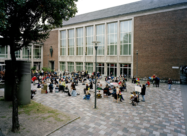 a moment of the reading performance at Theo-Burauen-Platz: many people sitting outside in front of a building